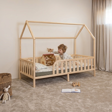 A young child stands inside a light natural wood house-shaped toddler bed in a softly lit nursery. The bed has a slatted guard rail, low side rails, and a roof frame.