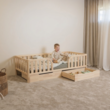 A toddler sits on a light natural wood toddler bed with built-in storage drawers, styled in a bright, neutral-toned nursery with beige carpet and curtains.