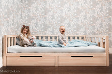 Wooden Montessori toddler bed with storage drawers and two children sitting on the mattress